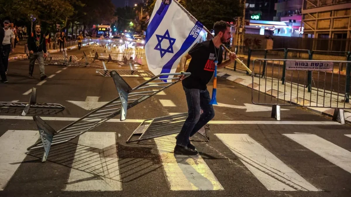 Seorang pengunjuk rasa merobohkan barikade sambil memegang bendera Israel selama unjuk rasa besar di Tel Aviv